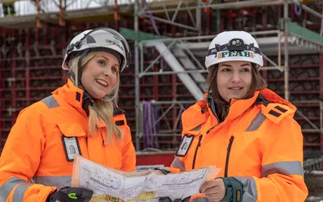 Two women on a building site with hard hats and orange coats with PEAB logo