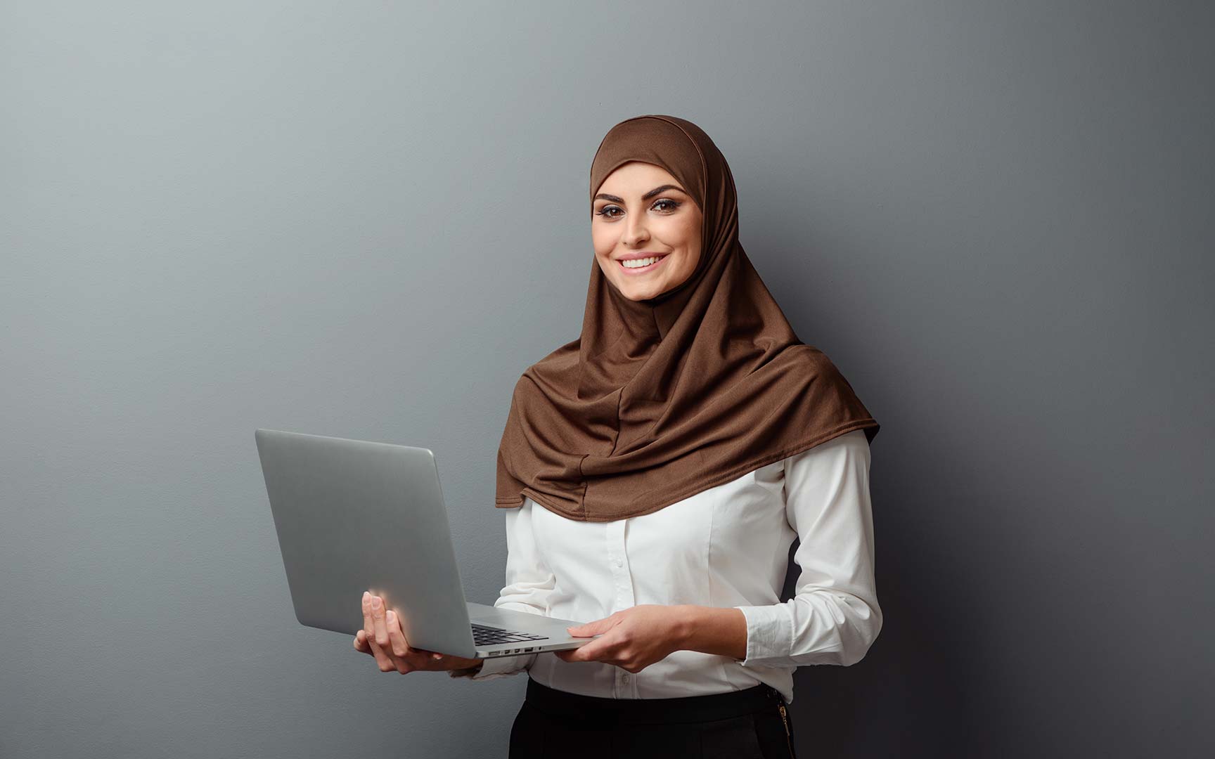 A woman wearing a brown hijab holds a laptop, smiling against a gray background. 