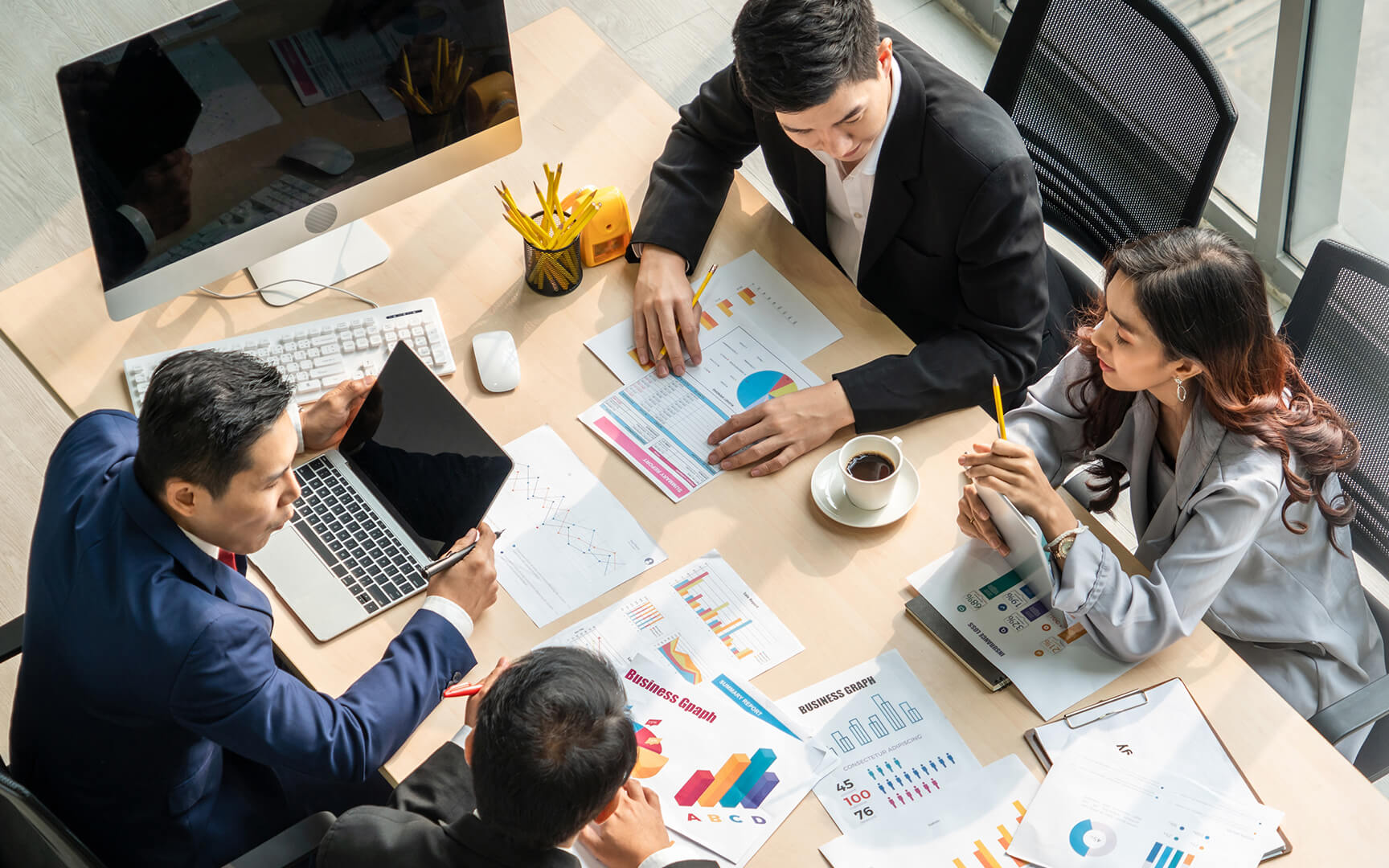 A group of four professionals discussing charts and data at a conference table. One person has a laptop