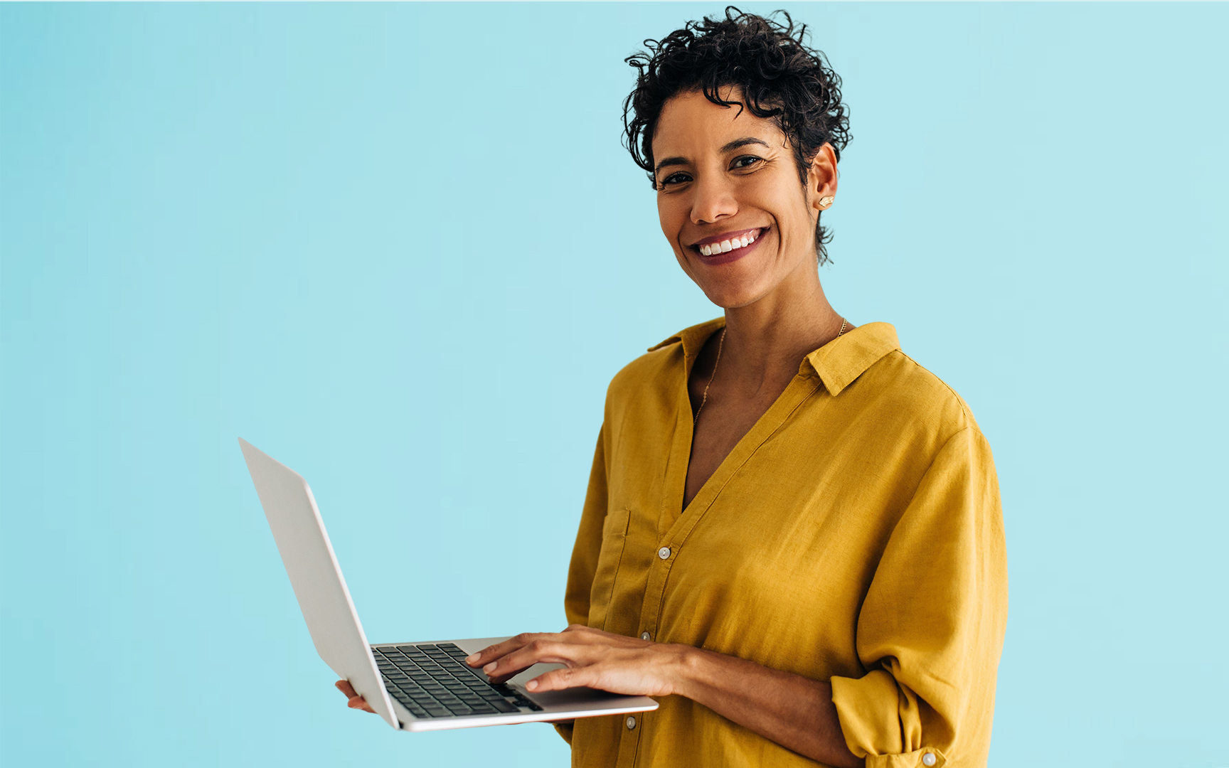 A woman with curly hair smiles while holding a laptop in front of a blue background