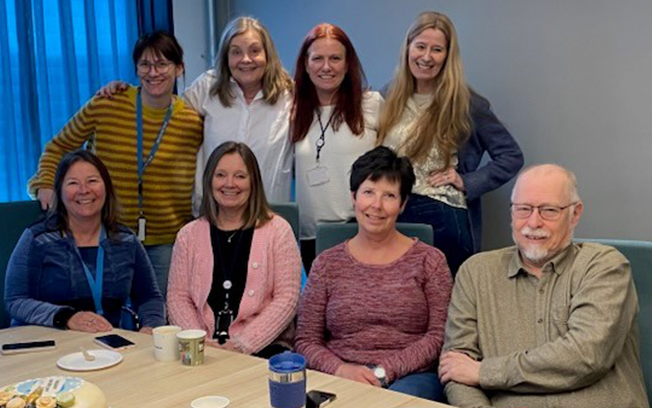 A group of eight people smiling around a table with coffee cups and a cake. 