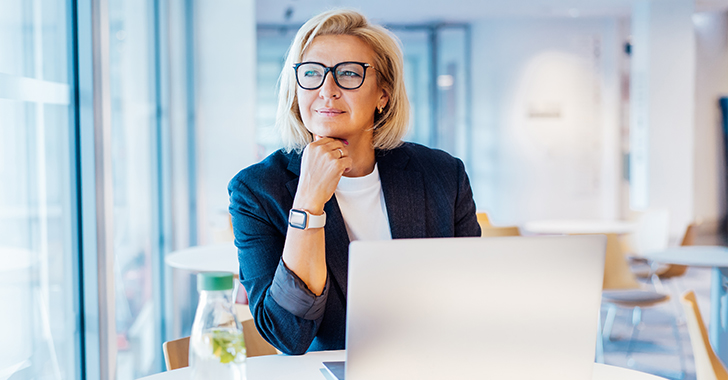 A thoughtful woman with blonde hair and glasses sits by a laptop in a modern workspace. 