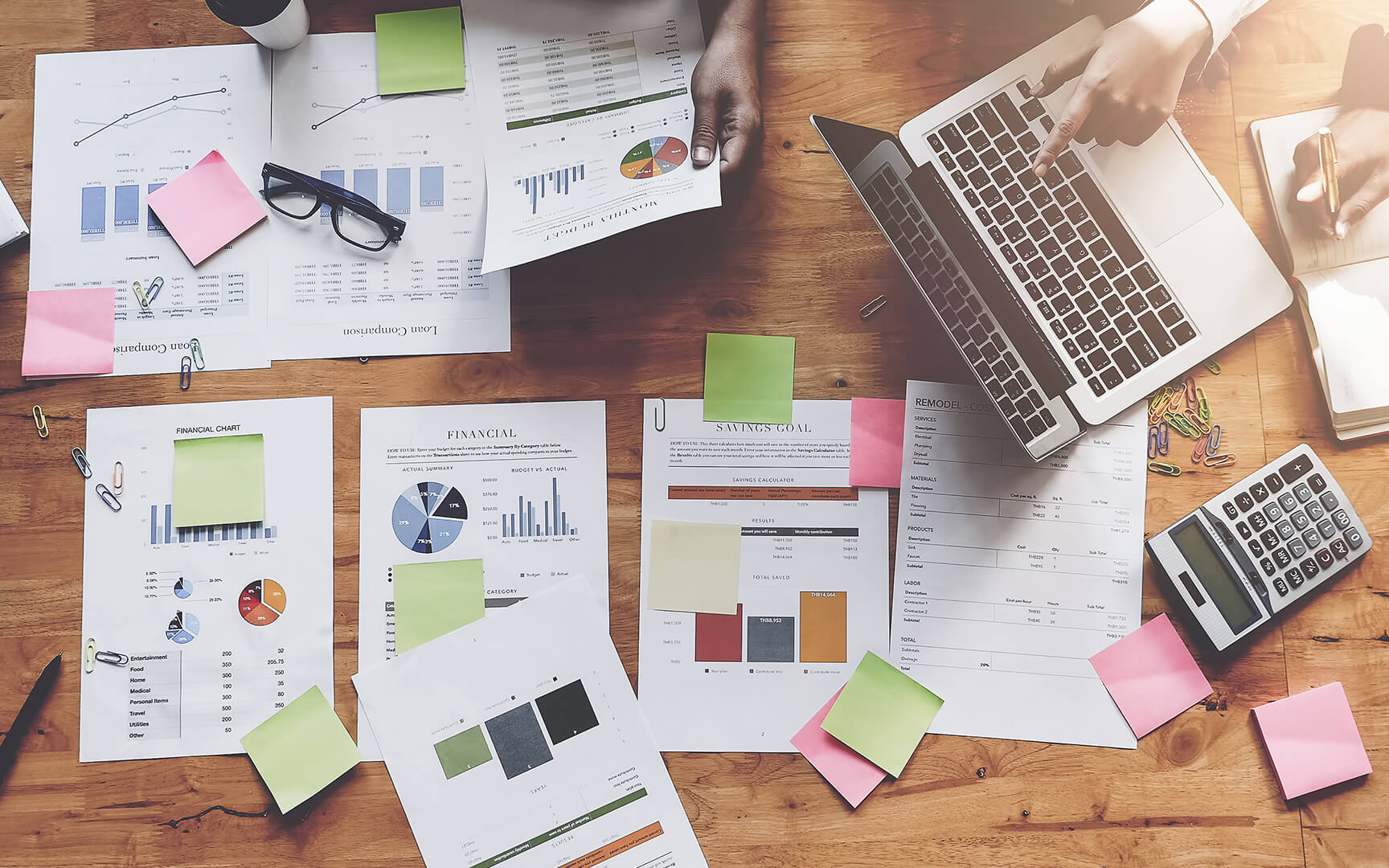 A wooden table with financial documents, a laptop, sticky notes, a calculator, and a notebook. 