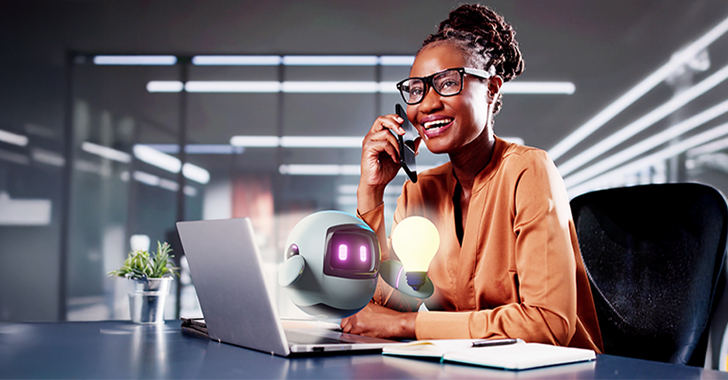 A woman smiling on a phone while sitting at a desk with a laptop and a cartoon robot holding a light bulb. 