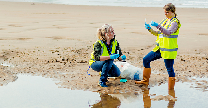 Two women in yellow vests collecting samples and cleaning the beach, with trash bags nearby.
