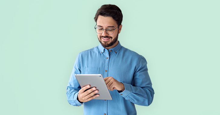 A man in a blue shirt smiles while using a tablet in front of a light green background. 