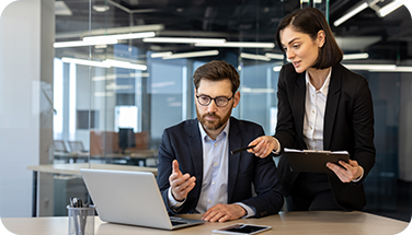 A man in glasses sits at a laptop while a woman in business attire stands beside him, pointing and discussing. 