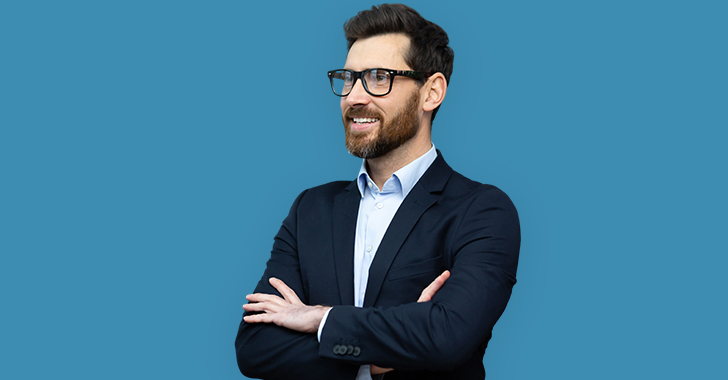 A man in a dark suit and glasses smiling, with arms crossed in front of a blue background. 