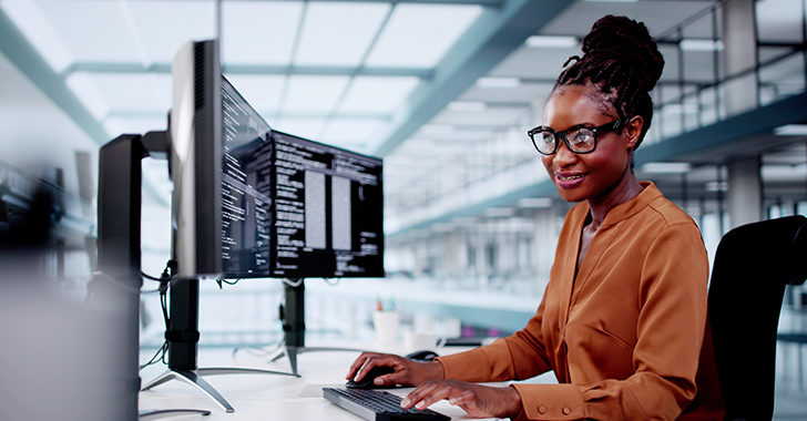 A professional seated at a desk in a modern office, working on a dual-monitor setup displaying code or text, wearing glasses and a brown shirt, smiling and engaged in their task.