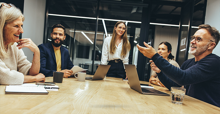 A group of five people in a meeting room, smiling and discussing at a table with laptops and drinks. 