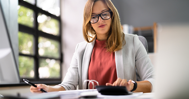 A professional woman with light brown hair and black-framed glasses works at a desk in a bright office.