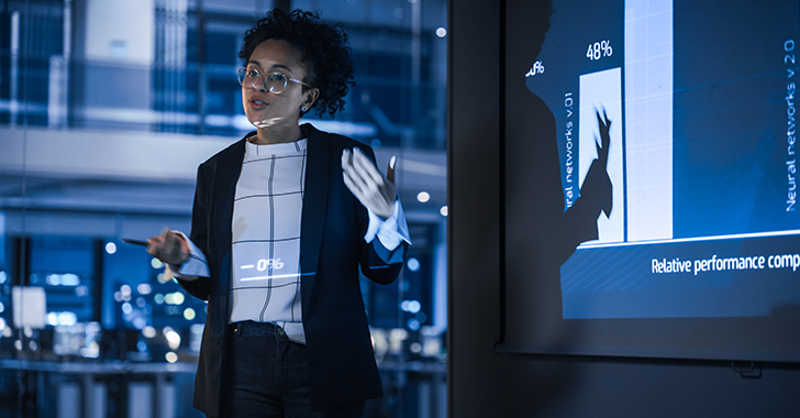 A professional presentation scene featuring a woman with dark curly hair and glasses, gesturing while explaining a bar chart projected on a screen.