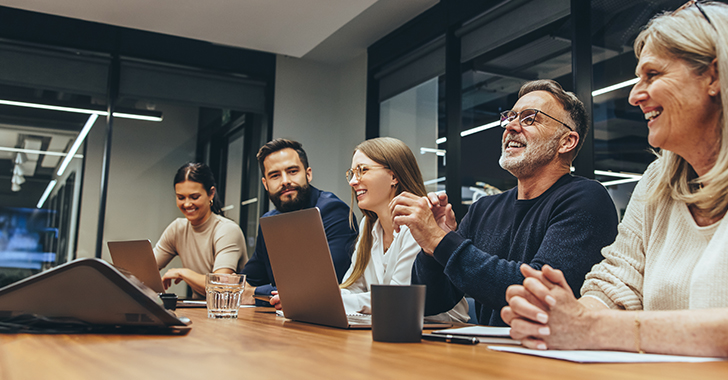 A modern office setting with five individuals seated around a wooden conference table, engaged in a collaborative meeting. 