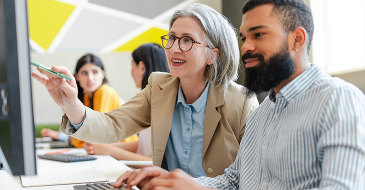 A professional setting with four individuals, where a woman with gray hair and glasses points at a computer screen with a pen, smiling. 