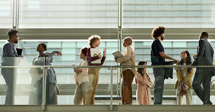 A diverse group of professionals interact on a modern, glass-railed walkway in a bright, naturally lit building. 