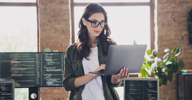 A young woman with long dark brown hair and glasses works intently on a silver laptop in a modern office.