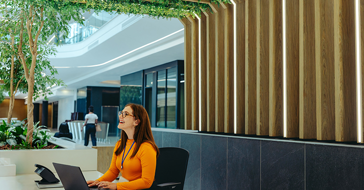 A smiling woman with reddish-brown hair and glasses sits at a white desk in a modern office lobby. 