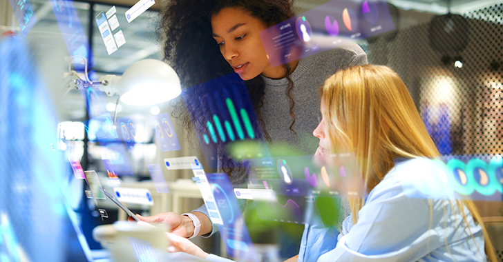 Two women collaborate in a modern office, surrounded by glowing holographic data visualizations.