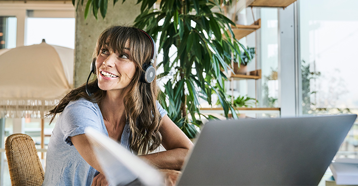 A smiling woman with wavy brown hair and headphones with a microphone sits at a desk with a silver laptop. 