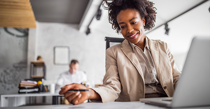 A woman in a beige blazer smiles while writing notes, with a laptop in front of her. 