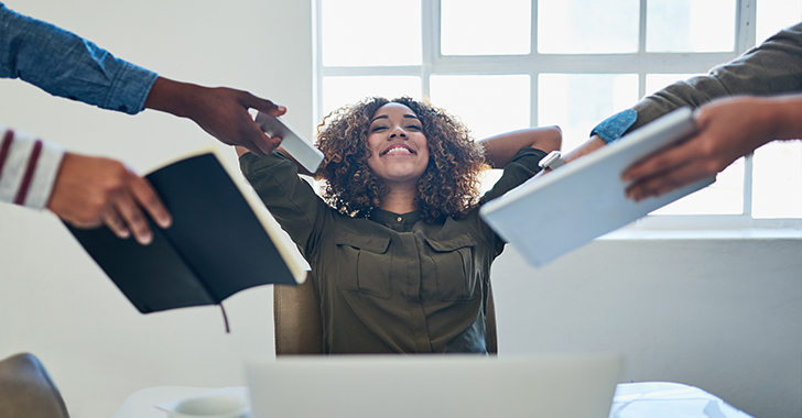 A smiling woman sitting at a desk with headphones, relaxing with hands behind her head while receiving books from others. 