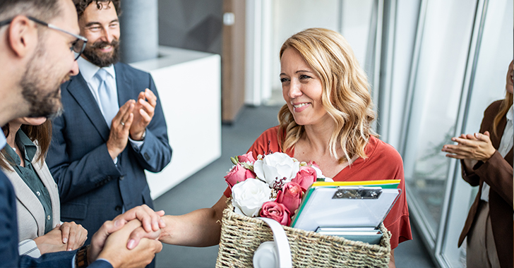 A blonde woman in a reddish-orange top smiles warmly as she shakes hands with a male colleague in a modern office hallway.