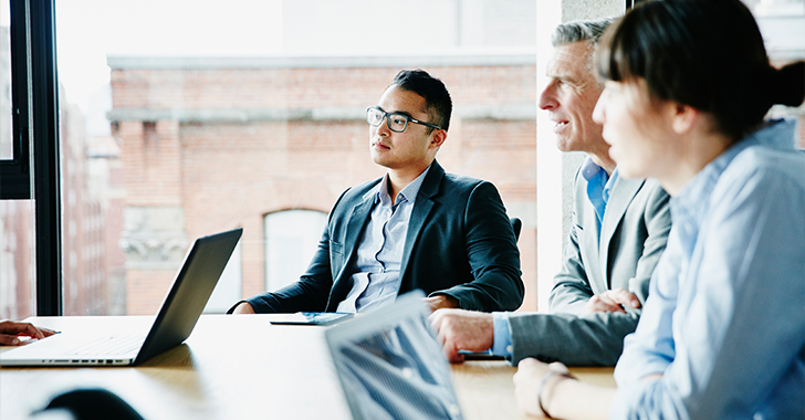 Three professionals sit around a wooden conference table in a modern office with large windows and exposed brick walls.