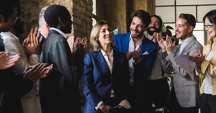 A group of people applauding a woman in a suit, celebrating a success in a bright room. 