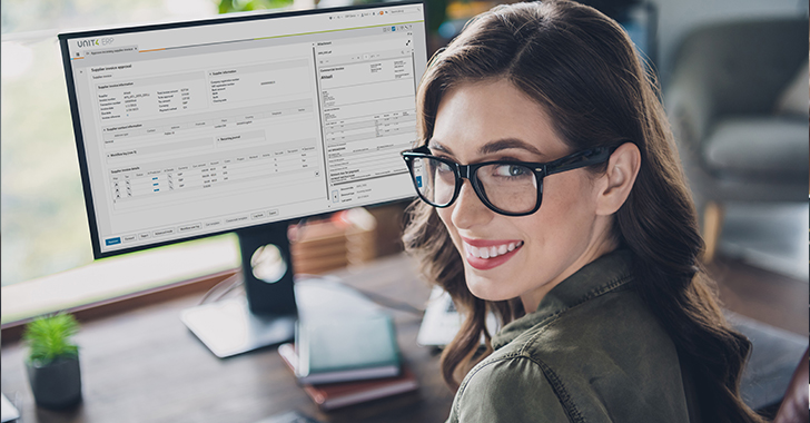A smiling woman in glasses looks over her shoulder at the camera while working at a desk.