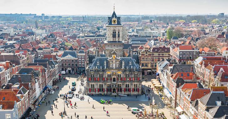 Aerial view of a historic town square with a large clock tower and red-roofed buildings. 