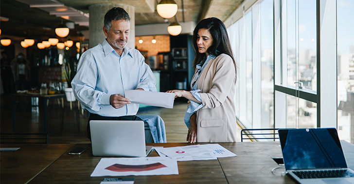 Two professionals discussing documents at a table with laptops and papers, in a modern office. 