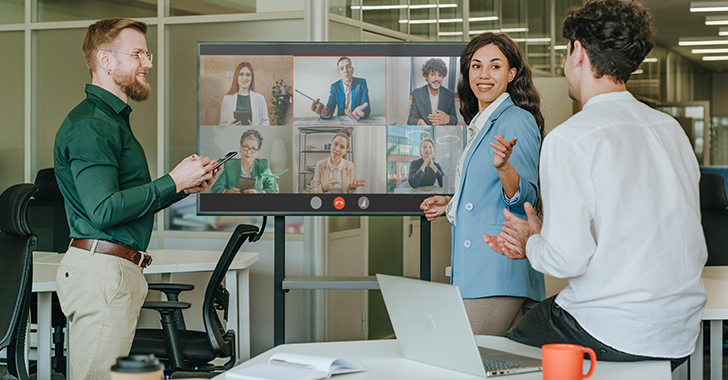 A group of six people in a modern office meeting, discussing at a large table with laptops and mugs. 