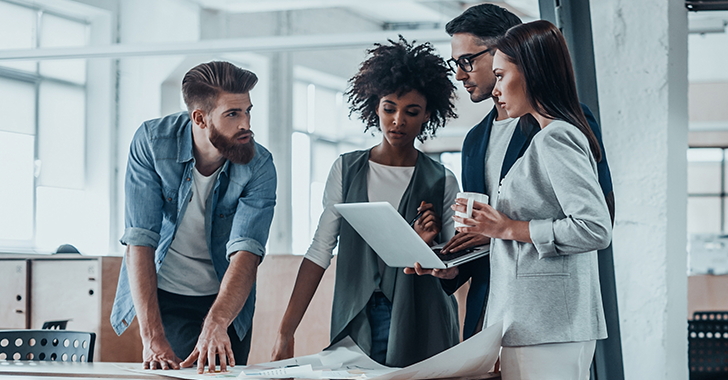 Four people in a modern office discussing a project on a laptop and paperwork. 