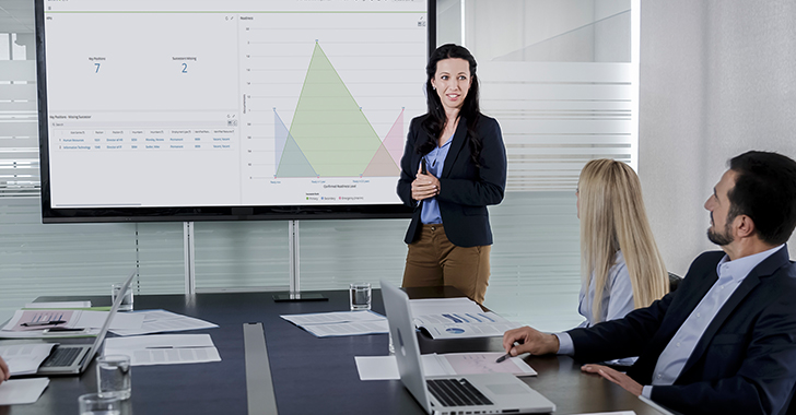 A woman presents data on a screen to a meeting room of people with laptops and papers. 