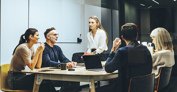 A team meeting in a modern office with six professionals at a table, discussing with a laptop and coffee cups. 