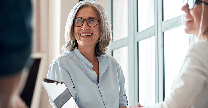 Two people are seated near large windows in a bright indoor setting, engaged in conversation.