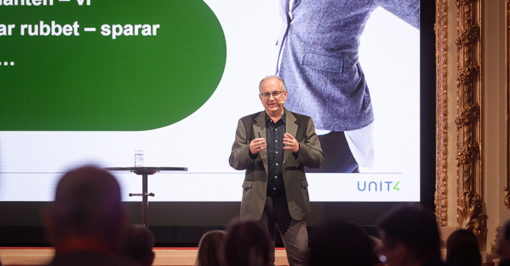 A speaker in a gray suit presents on stage in front of a large screen. 