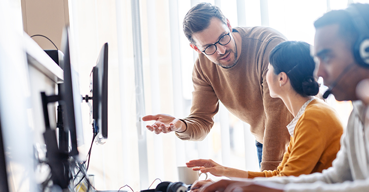 A person standing beside a seated colleague in an office setting, gesturing toward a computer screen while discussing work. 
