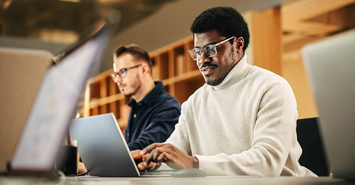 Several people are seated at a long desk in a modern, warm‑toned workspace, working on laptop computers. 