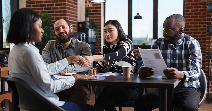 A diverse group of professionals seated around a table in a modern office, reviewing printed documents and exchanging papers during a collaborative meeting, with brick walls, large windows, and pendant lighting in the background.