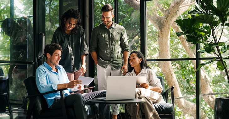 A group of four people are gathered in a modern office space with large windows and abundant natural light. 
