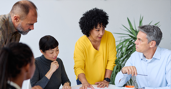 A group of five people gathered around a table in a bright meeting room, reviewing documents and discussing work. 