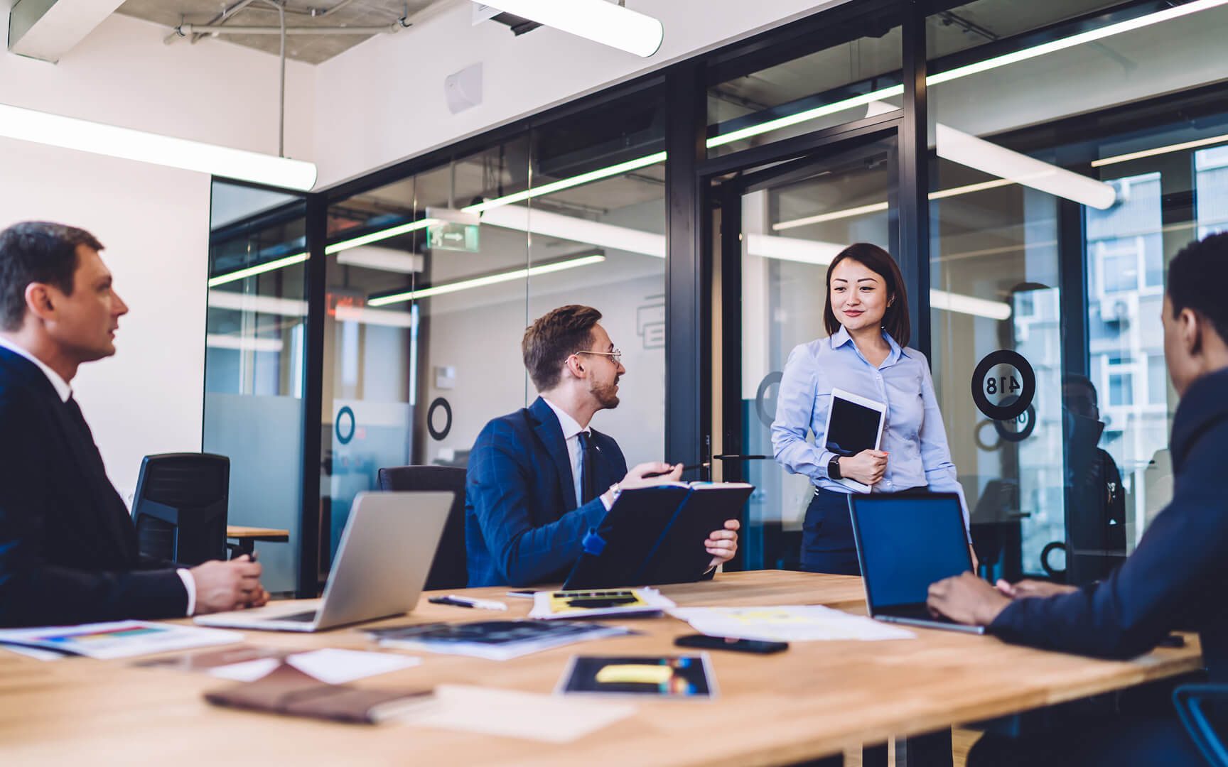 A group of professionals collaborating in a modern office meeting room, seated around a table with laptops and documents, while one colleague stands and presents ideas near glass walls and large windows.