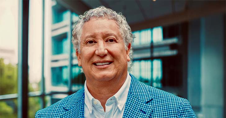 Portrait of a professional wearing a blue patterned blazer and white shirt, standing indoors in a modern office environment with glass walls and natural light in the background.