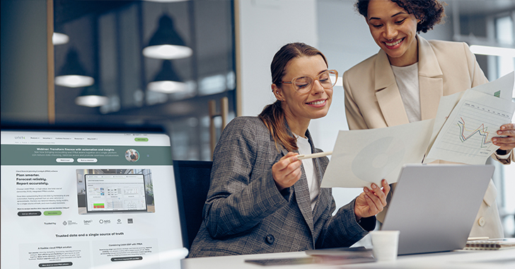 Two professionals collaborating at a desk in a modern office, reviewing printed reports and digital dashboards on a laptop and desktop screen.