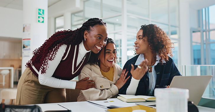Three people collaborate at a table in a bright, modern learning space, discussing notes and ideas while working with a laptop, notebooks, and documents, suggesting teamwork and academic or professional collaboration.
