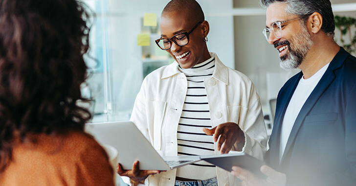 Three professionals stand in a modern office environment, reviewing information on a tablet together while discussing a document, suggesting collaboration and decision‑making in a workplace setting.