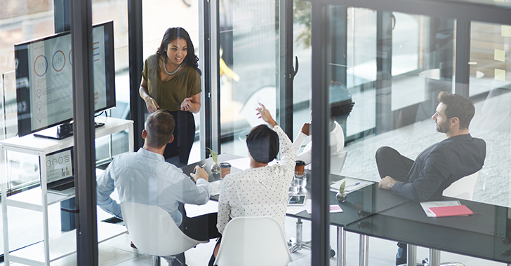 Groupe de collaborateurs dans une salle de réunion vitrée discutant de données affichées sur un écran de présentation.