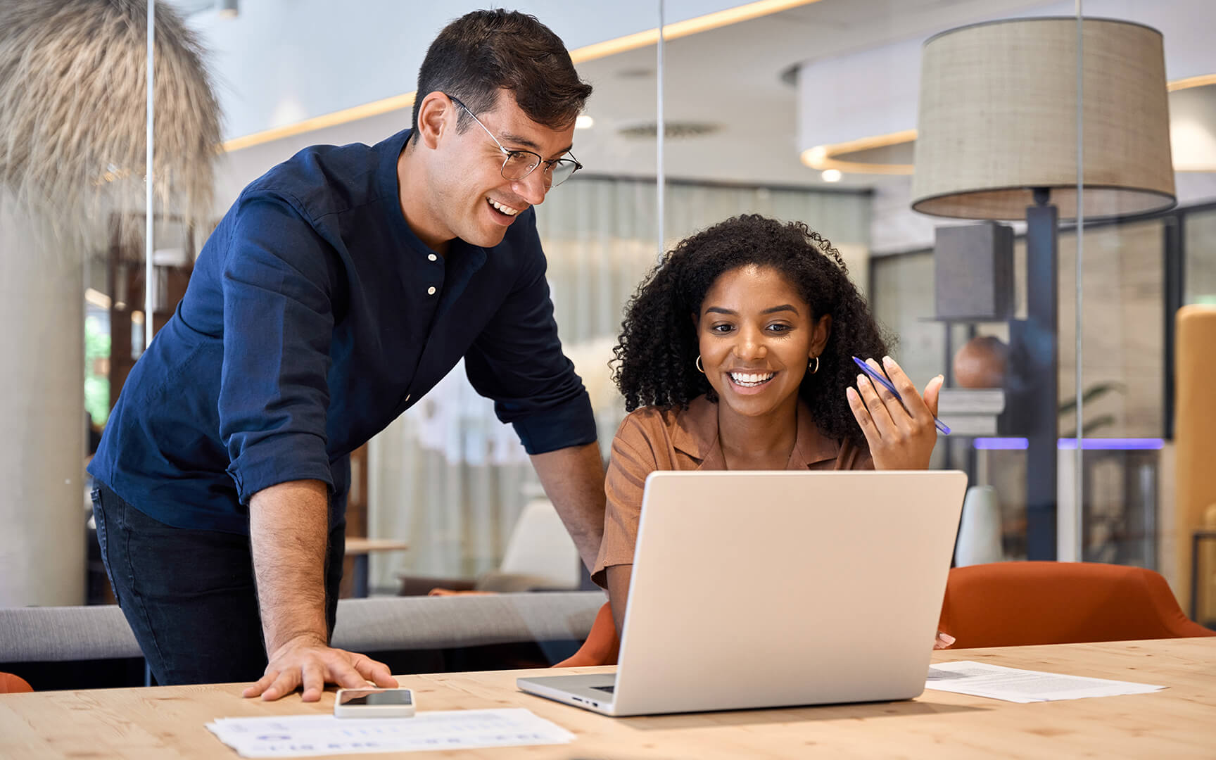 Två kollegor samarbetar vid ett skrivbord på ett modernt kontor, granskar dokument och diskuterar insikter framför en bärbar dator i en ljus arbetsmiljö.