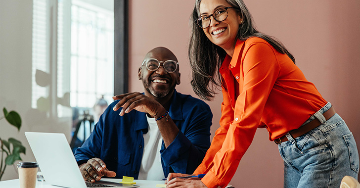Two colleagues working at a desk with a laptop, reviewing documents together in a modern office environment.
